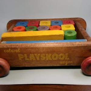 May include: A vintage wooden Playskool wagon with colorful blocks. The wagon is made of wood and has red wheels. The blocks are various colors, including red, blue, yellow, and green. The text "Playskool wagon" is printed on the side.