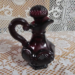 May include: A dark red glass pitcher with a decorative design and a knobbed lid. The pitcher has a curved handle and a wide base.