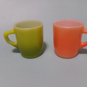 May include: Two small glass mugs with a smooth finish and a ribbed base. One mug is a vibrant lime green color, while the other mug is a bright orange color. Both mugs feature a sturdy, angular handle attached to the side of the cylindrical body.