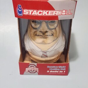 May include: A StackerDoll Russian-style nesting doll set featuring 5 dolls in 1, packaged in a red and white box. The box displays the text Legends of the Shoe, StackerDoll, and Ohio State. The doll inside wears glasses, a white shirt, and a tie.