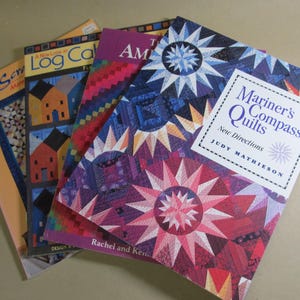 May include: A stack of quilting books with colorful covers. The titles include "Mariner's Compass Quilts," "A New Look at Log Cabin," and "Some". The covers feature quilt patterns with star and geometric designs, in shades of blue, red, purple, and orange.