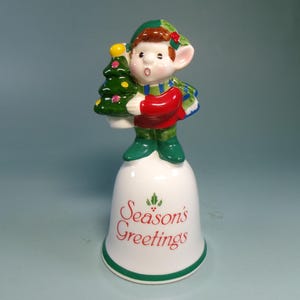 May include: A ceramic Christmas bell featuring a figurine of an elf holding a green Christmas tree. The elf wears a red shirt, green shoes, and a green and white scarf. The bell is white with "Season's Greetings" in red.