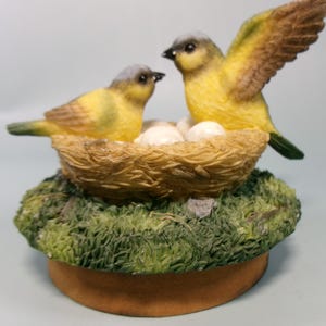 Peut inclure: Figurine d&eacute;corative repr&eacute;sentant deux oiseaux jaunes avec des t&ecirc;tes grises et des ailes marron. L'un des oiseaux a son aile d&eacute;ploy&eacute;e. Ils sont pr&egrave;s d'un nid avec des &oelig;ufs, sur une base moussue et une plateforme circulaire brune.