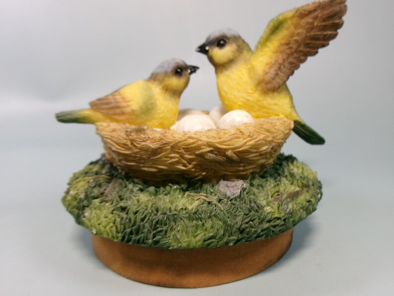 Peut inclure: Figurine d&eacute;corative repr&eacute;sentant deux oiseaux jaunes avec des t&ecirc;tes grises et des ailes marron. L'un des oiseaux a son aile d&eacute;ploy&eacute;e. Ils sont pr&egrave;s d'un nid avec des &oelig;ufs, sur une base moussue et une plateforme circulaire brune.