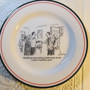 May include: A white ceramic plate with a black rim and a red band around the edge. The plate features a black and white cartoon illustration of people in a room with the caption "We didn't have time to pick up a bottle of wine, but this is what we would have spent."