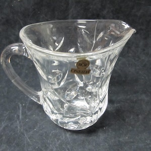 May include: A clear glass creamer with a cut glass design. The creamer has a handle and a label that reads "RCR Oneida".