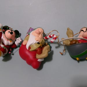 May include: Three holiday ornaments featuring Disney characters: Mickey Mouse dressed as Santa tangled in string lights, a dwarf with a white beard holding a candy cane and a red sack, and a dwarf sitting in a gray cauldron with a green bow.