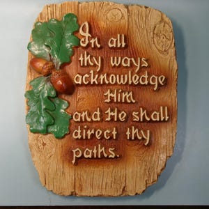 May include: A rectangular wooden plaque with the quote "In all thy ways acknowledge Him and He shall direct thy paths." The plaque features green oak leaves and brown acorns. The background is a light brown with wood grain.