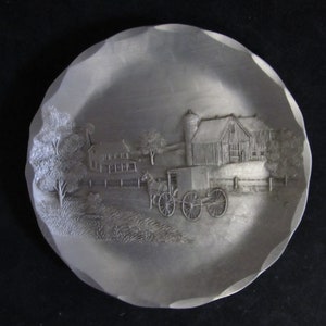 May include: A silver-toned metal plate with a raised design of a farmhouse with a barn, a horse-drawn buggy, and trees. The plate has a scalloped edge.