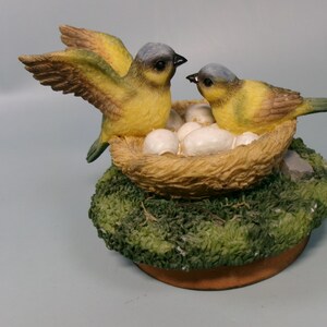 Peut inclure: Figurine d&eacute;corative repr&eacute;sentant deux oiseaux jaunes avec des t&ecirc;tes grises et des becs noirs. L'un des oiseaux a les ailes d&eacute;ploy&eacute;es, l'autre est assis &agrave; c&ocirc;t&eacute; d'un nid rempli d'&oelig;ufs blancs. Le nid est pos&eacute; sur un socle mousseux.