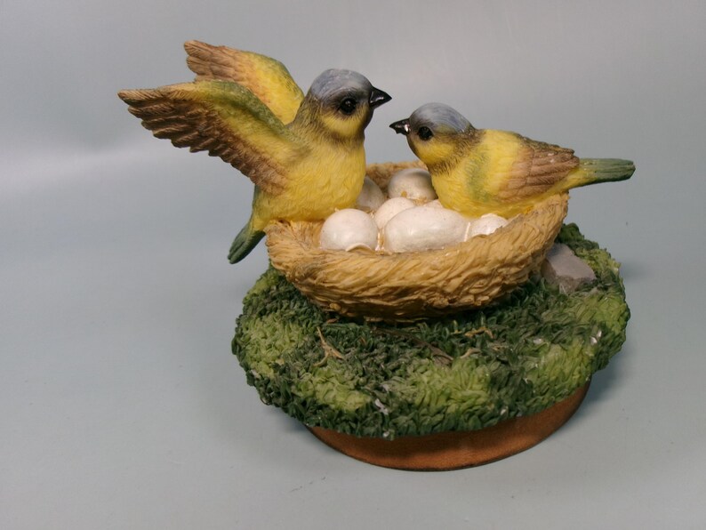 Peut inclure: Figurine d&eacute;corative repr&eacute;sentant deux oiseaux jaunes avec des t&ecirc;tes grises et des becs noirs. L'un des oiseaux a les ailes d&eacute;ploy&eacute;es, l'autre est assis &agrave; c&ocirc;t&eacute; d'un nid rempli d'&oelig;ufs blancs. Le nid est pos&eacute; sur un socle mousseux.
