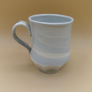 May include: A ceramic mug featuring a marbled pattern of soft blue and white swirls. The mug has a curved handle on the left side and a slightly tapered body that widens toward the rim, with a smooth, glossy finish throughout the exterior surface.