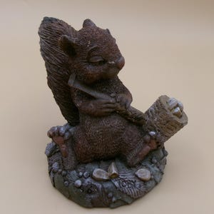May include: A brown squirrel figurine sitting on a textured base with scattered nuts. The squirrel holds a long, thin tool in its paws and is positioned next to a small, cylindrical basket filled with several light-colored nuts or seeds.