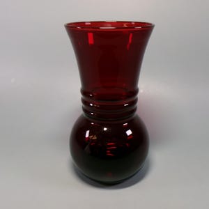 May include: A ruby red glass vase with a bulbous base and a flared top. The vase has a series of horizontal ridges around the middle. The glass is transparent, allowing light to pass through, and has a glossy finish. The vase is likely used for decorative purposes.