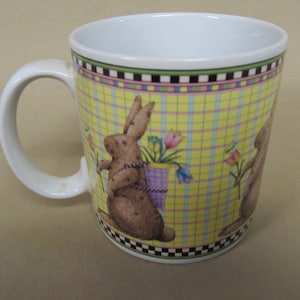 May include: White ceramic mug with a yellow, pink, and blue plaid pattern. The mug features a brown rabbit carrying a basket of flowers.