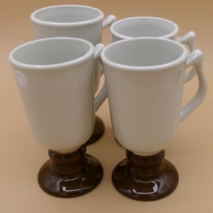 May include: A set of four ceramic mugs, each featuring a glossy white upper body and a contrasting dark brown pedestal base. The mugs have a tall, tapered shape with a curved handle on the side, designed for serving hot beverages in a footed style.
