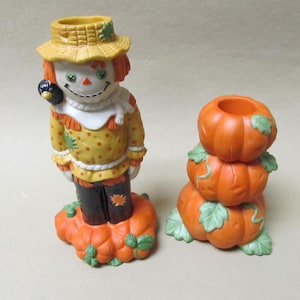May include: Two ceramic Halloween decorations. One is a scarecrow figurine with a yellow hat, orange hair, and a black bird on its shoulder. The other is a stack of three orange pumpkin candle holders with green leaves.