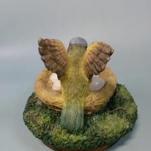 Peut inclure: Figurine d'oiseau aux ailes d&eacute;ploy&eacute;es, au-dessus d'un nid avec des &oelig;ufs. L'oiseau a des plumes jaunes et vertes, avec des ailes marron. Le nid est fait de paille et repose sur une base verte et moussue.