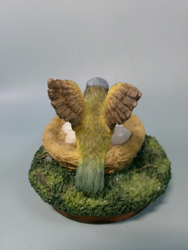 Peut inclure: Figurine d'oiseau aux ailes d&eacute;ploy&eacute;es, au-dessus d'un nid avec des &oelig;ufs. L'oiseau a des plumes jaunes et vertes, avec des ailes marron. Le nid est fait de paille et repose sur une base verte et moussue.