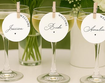 May include: Three champagne glasses with name tags that read "Jessica", "Bride", and "Amelia". Each glass is filled with a light yellow liquid and is set on a white table. White flowers are in the background.