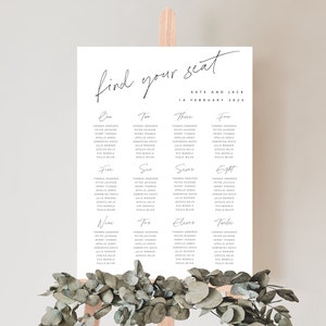 May include: A white wedding seating chart with the text "find your seat" in black cursive script. The chart lists table numbers from one to twelve with guest names listed under each table number. The chart is on an easel with greenery in front of it.