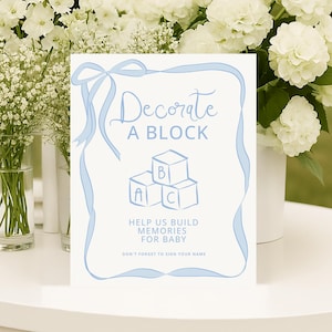 May include: A white sign with blue text and a ribbon design, reading "Decorate A Block" with block illustrations. The sign is surrounded by white flowers in vases, perfect for a baby shower or event.