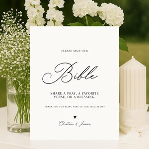 May include: A white sign with the word "Bible" in elegant script, surrounded by white flowers and a candle. The sign reads "Please Sign Our Bible" and invites guests to share a prayer, verse, or blessing. The names "Christian & Jessica" are at the bottom.