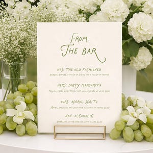 Peut inclure: Un menu de bar sur une table blanche, soutenu par un support en métal doré, avec des fleurs blanches et des raisins verts. Le menu propose des cocktails comme Old Fashioned, Dirty Margarita et Aperol Spritz, ainsi qu'une option sans alcool.