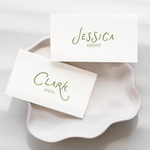 May include: Two white place cards with green calligraphy names "Jessica Roberts" and "Clark Smith" rest on a white, scalloped-edge dish. The cards are arranged on a light gray surface, with shadows.