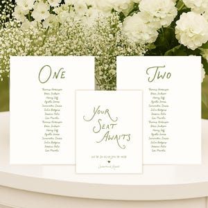 May include: Wedding table plan with white cards and green lettering. The cards are in front of white flowers. The cards read "ONE" and "TWO" with guest names, and "YOUR SEAT AWAITS".