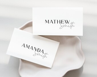 May include: Two white business cards with names printed in black, "MATHEW Smith" and "AMANDA Smith", rest on a white, scalloped-edge dish. The names are in a combination of serif and script fonts. The background is a soft, neutral tone.