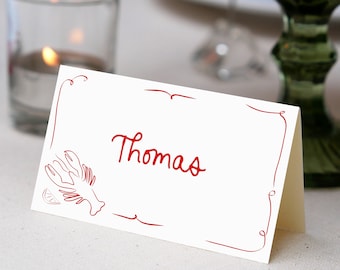 May include: A white place card with a red border and a lobster illustration. The name "Thomas" is written in red cursive.