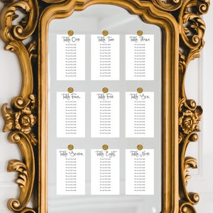 May include: A gold-framed mirror with nine white cards attached to it. Each card has a gold circle at the top and lists the names of guests assigned to each table. The cards are labeled "Table One", "Table Two", "Table Three", "Table Four", "Table Five", "Table Six", "Table Seven", "Table Eight", and "Table Nine".