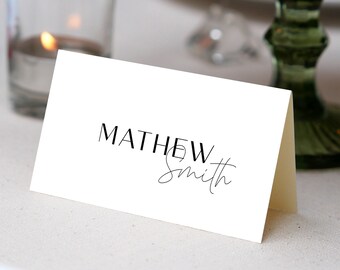 May include: White place card with black text that reads "MATHEW Smith" in a modern font. The card is folded in half and sitting on a white table.
