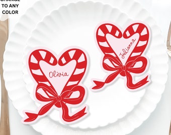 May include: Two red and white candy cane heart-shaped place cards with a red bow. The place cards are on a white plate with the names "Olivia" and "Julianna" written on them. The place cards are approximately 5x6 inches.