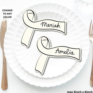May include: Two white ribbon place card holders with black calligraphy names "Mariah" and "Amelia". The place cards are on a white plate with a scalloped edge. The plate is 5 inches by 6 inches.