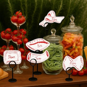 May include: A wooden table displays various food items and decorative elements. Cherry tomatoes are in glass goblets and a wooden bowl. Pasta is in glass jars. Numbered signs in red and white are on black stands. Greenery is in the background.