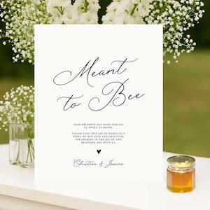 May include: A wedding sign with the text "Meant to Bee" in elegant script, accompanied by a small jar of honey. White flowers and greenery frame the sign, creating a romantic and natural aesthetic.