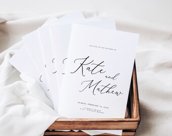 Può includere: Una pila di programmi di nozze bianchi con testo di calligrafia nera. I programmi sono in un vassoio di legno e presentano i nomi "Kate e Matthew" e la data del matrimonio.
