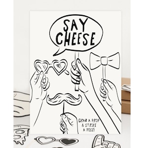 Può includere: Illustrazione in bianco e nero di un set di accessori per fotomaton con il testo "Say Cheese". Il set include un papillon, un baffo, occhiali da sole e un cartello che dice "Grab a prop & strike a pose!"