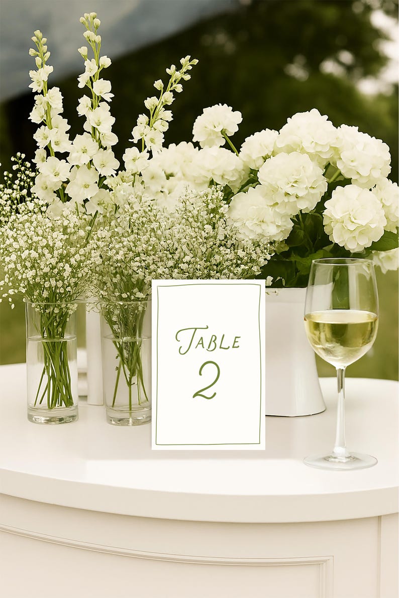May include: A white table setting with floral arrangements and a wine glass. The table features a sign that reads "Table 2" in green script. The flowers include white hydrangeas, baby's breath, and other white blooms.