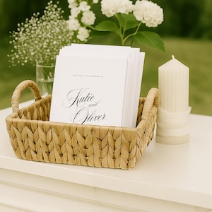 May include: A rectangular woven basket with two handles containing a stack of white wedding programs. The programs feature the text "Welcome to the wedding of Katie and Oliver" in black script and serif font. A ribbed white pillar candle sits nearby.