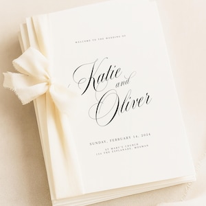 以下が含まれることがあります： クリーム色のリボンで結ばれた、白いウェディングプログラムの束。「Welcome to the Wedding of Katie and Oliver」と日付と場所が印刷されています。