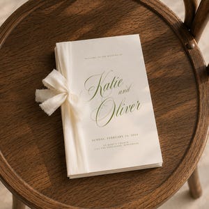 May include: A white wedding program booklet with green text reading "Welcome to the wedding of Katie and Oliver, Sunday, February 14, 2024, St Mary's Church, 1234 The Esplanade, Kingshead." The booklet is bound with a cream-colored frayed fabric ribbon.