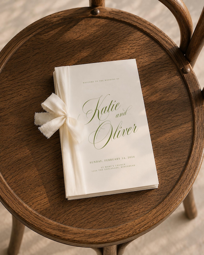 May include: A white wedding program booklet with green text reading "Welcome to the wedding of Katie and Oliver, Sunday, February 14, 2024, St Mary's Church, 1234 The Esplanade, Kingshead." The booklet is bound with a cream-colored frayed fabric ribbon.