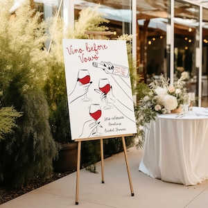 May include: A white sign with black line drawings of wine glasses and a bottle of wine. The sign says "Vino before Vows" and "Let's celebrate Amelia's Bridal Shower". The sign is on a wooden easel.