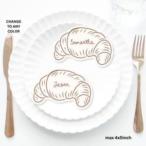 Op de afbeelding: Twee croissantvormige naambordjes, één met de naam "Samantha" en de andere met "Jason", liggen op een wit bord. De kaarten zijn bruin met een crèmekleurige achtergrond. Aan weerszijden van het bord liggen een mes en een vork. De tekst "CHANGE TO ANY COLOR" staat linksboven.