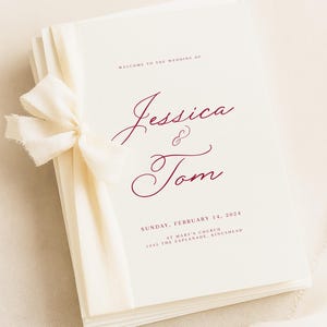 Può includere: Un libretto per il programma di matrimonio color crema con testo in corsivo rosso scuro che recita Jessica & Tom, insieme alla data domenica 14 febbraio 2024 e alla posizione St. Mary's Church, 1345 The Esplanade, Kingshead, legato con un nastro crema.