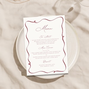 Puede incluir: Una tarjeta de menú blanca con un borde burdeos descansa sobre un plato blanco. El menú enumera los platos: Para empezar, Plato principal y Postre. El menú incluye pan artesanal, ricotta batida y paletilla de cordero cocida a fuego lento.