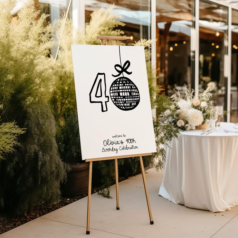 Op de afbeelding: Een wit bord met zwarte tekst en een zwart-witte discobal-afbeelding. Het bord staat: "Welkom op Olivia's 40e verjaardagviering". Het bord staat op een ezel.